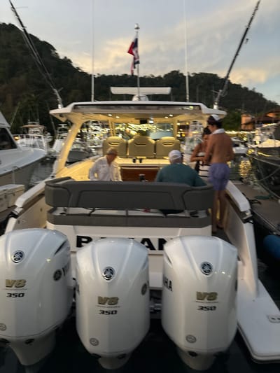 The image shows the rear of a white motorboat docked in a marina in Playa Herradura, Costa Rica. Three people are on board the boat. One man is wearing a white shirt and is looking at a cell phone in his hand. Another man is wearing a dark blue shirt and is looking off to the right. A third man is shirtless, wearing white shorts, and is looking to his left. The boat has three outboard motors, each with the words "V8 350" printed on them. The boat is docked alongside several other boats. There are lush green trees on the shore behind the boats, and the sky is a pale blue with a hint of orange. There is a small American flag flying from the top of the boat.