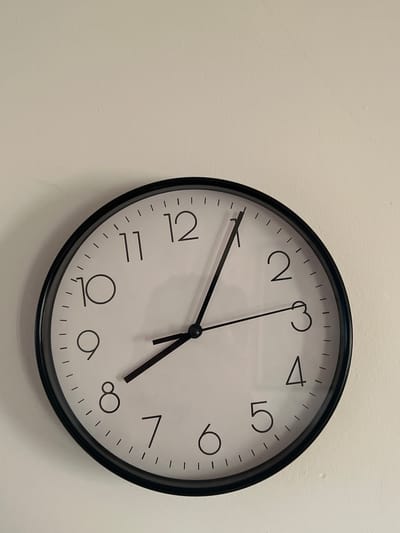 This media file is an image of a black-framed analog clock mounted on a light-colored wall. The clock face is white with black numerals and hour markers. The hour hand is pointing just before the 5, and the minute hand is pointing at the 4. This indicates the time is approximately 4:23. The background is a plain, textured wall, with no other objects or figures visible. There is no visible text in the image, except for the numerals on the clock face. The lighting suggests an indoor setting, possibly during daylight hours. The image focuses solely on the clock and its immediate surroundings.