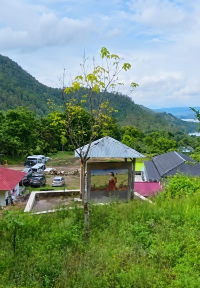 In Sosor Dolok, Indonesia, a scenic overview captures a cluster of buildings nestled amidst lush green hills. A young tree with bright green leaves stands in the foreground, a stark contrast to the overcast sky. To the left, a white van and several cars are parked near a building with a red roof. A mural depicting two figures in a field adorns a small structure with a corrugated metal roof, positioned centrally in the frame. In the background, a vast expanse of verdant hills stretches towards a distant, hazy body of water, hinting at a serene natural landscape.