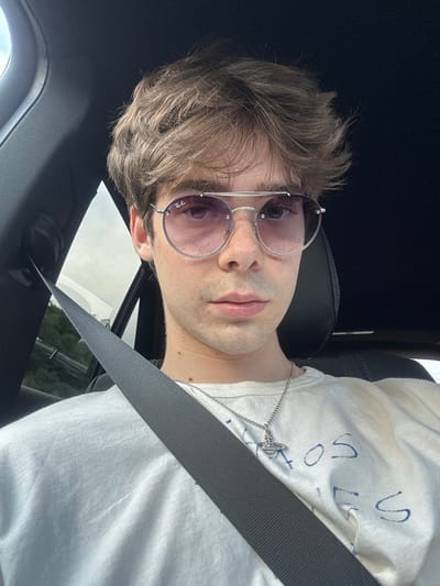 A young Caucasian male with light brown, tousled hair and freckles across his nose and cheeks is pictured from the chest up, looking directly at the camera. He is wearing large, round, silver-rimmed sunglasses with pink-tinted lenses and a white t-shirt with blue writing. A silver chain with a Saturn pendant is visible around his neck. He is seated in the driver's seat of a vehicle, with a grey seatbelt diagonally across his chest. The interior of the car is visible, with dark upholstery and a glimpse of greenery and a cloudy sky through the window on the left side. The scene appears to be set on a road, possibly on the outskirts of Bela Cerkev, Slovenia, given the visible vegetation and highway infrastructure in the background.
