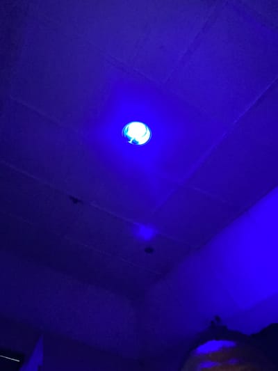 The image shows a dimly lit interior space, likely a room, with a predominantly blue hue cast by artificial lighting. The ceiling appears to be made of modular tiles, with subtle lines indicating seams. A bright blue light source is visible in the upper central portion of the image, appearing to be a light fixture or LED. There is a faint, circular shadow or light reflection on the ceiling below the main light source. Towards the bottom of the frame, there are indistinct shapes and colors, suggesting the presence of objects or people, possibly a face with illuminated features, though this is unclear due to the low light and color cast. The overall impression is of a low-light environment with prominent blue illumination. The location is specified as Edopa, Nigeria.