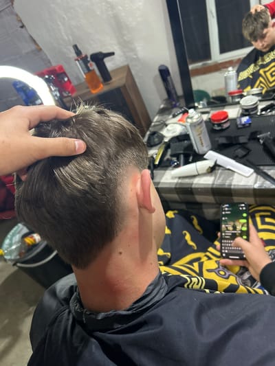 This image depicts an indoor grooming establishment, likely a barbershop, located in Hvostiane, Bulgaria.

In the foreground, a young male is seated, facing away from the camera. He wears a dark blue or black barber's cape, secured around his neck. His short, brown hair shows a faded cut on the sides and back, with longer hair on top, which is being held by a visible left hand from an unseen individual, presumably the barber. A reddish patch is visible on the client's lower neck. The client's right hand holds a smartphone, its screen displaying a chat application with text and images.

To the right, a checkered tablecloth covers a counter, upon which various barber tools and products are arranged. These include multiple hair clippers/trimmers, hair spray cans (one white, one orange), jars of hair styling products, brushes, combs (one white), and additional spray bottles. A large mirror dominates the background, reflecting the interior. In the mirror's reflection, another young male, possibly a different client, is visible wearing a yellow and black geometric patterned barber's cape, looking downwards. Behind the reflected individual, a window is discernible.

On the left side of the image, a large circular LED ring light provides illumination. A dark wooden cabinet or shelf is partially visible behind the ring light, with an orange spray bottle and a black hairdryer or similar tool placed on it. A black bucket or bin is partially visible on the far left, near the floor. The walls of the establishment are light-colored and textured.