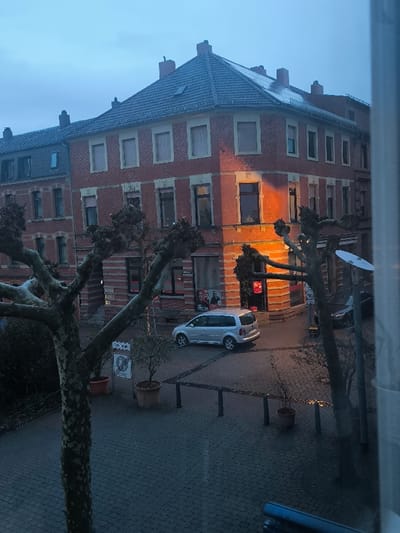 The image captures an elevated view of a street scene in Neunkirchen, Germany, under overcast conditions, likely at dusk or dawn. The central focus is a three-story red brick corner building with a dark tiled roof and multiple windows, some fitted with light-colored external blinds. A prominent orange light source illuminates a corner window and adjacent wall of this building. The ground floor of the corner building features commercial spaces with large display windows, one containing a poster depicting two individuals, and another faintly revealing a person inside behind a parked vehicle.

To the left, an adjacent multi-story red brick building with a dark roof continues the urban architectural style. The street below is paved with dark cobblestones, appearing damp or wet, reflecting ambient light. A silver Volkswagen minivan, likely a Touran, is parked on the cobblestone street near the corner building. The front section of a dark vehicle is partially visible on the far right.

Several large, heavily pruned deciduous trees with bare, gnarled branches dominate the foreground and frame the view. Potted plants are positioned on the cobblestone sidewalk. Black bollards define the edge of the pedestrian area, and a white traffic sign indicating a "ZONE" is visible. A white satellite dish is mounted on a pole or tree structure on the right side of the image. The sky is a uniform, light blue-grey. A window frame is partially visible on the extreme right, indicating the elevated vantage point.