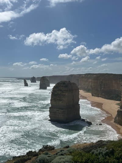 A scenic view of the Twelve Apostles, a collection of limestone stacks off the shore. The stacks rise from the ocean, with waves crashing around their bases. A sandy beach lines the coast, backed by tall cliffs. Vegetation covers the cliff tops on the right side. The sky is blue with scattered white clouds. The perspective is angled, tilting the horizon slightly.