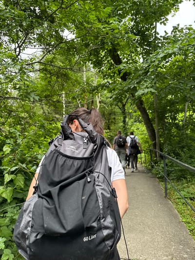 In Khleborob, Russia, a woman with dark hair pulled into a ponytail walks away from the camera on a paved path, wearing a large, black "60 Liter" backpack. She is dressed in a white t-shirt. Following her on the path are three men, also wearing backpacks and casual clothing. They are walking through a lush, green forest with dense foliage and trees lining the path, which is bordered by a black metal railing on the right side. The scene depicts a group hiking or walking in a natural, wooded area.