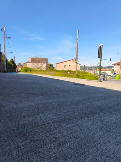 The scene depicts a street in Cangas de Morrazo, Spain, under a clear blue sky. The road surface is rough, marked with linear striations. Two pink-colored houses stand on the right side of the road, with grassy areas in front of them. Utility poles and wires line the roadside. A traffic light is visible, along with garbage bins. Other houses can be seen in the background.