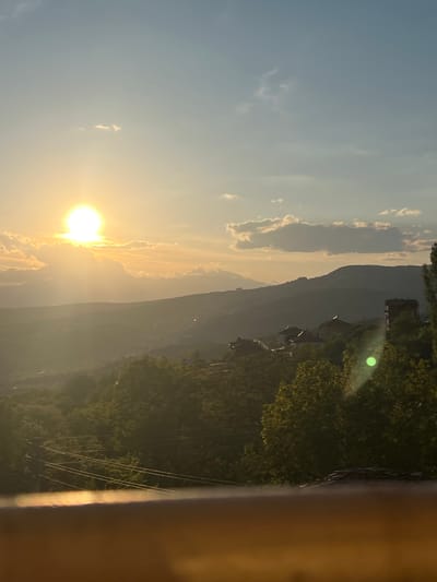 The image presents a landscape scene at sunset, viewed from an elevated position, likely a balcony or window. The sun is low in the sky, positioned behind a mountain range, and creating a bright, golden light that washes across the scene. Some clouds are visible. The surrounding terrain features a dense, tree-covered hillside with powerlines running across it, indicating a nearby settlement. Small structures, possibly houses, are visible, suggesting the outskirts of a residential area. Based on the location, the city of Kribul in Bulgaria, the landscape is characterized by hilly terrain and forest vegetation. The photograph suggests a calm, natural environment with the sun setting behind mountainous terrain.