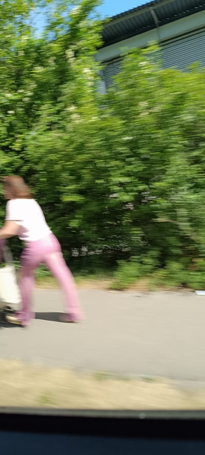 An individual, appearing to be a woman with shoulder-length brown hair, is captured in motion, pushing a wheeled shopping cart. She is wearing a light-colored t-shirt and loose, pink pants. The scene is blurred, indicating movement, likely from a vehicle. The setting appears to be a paved path next to a line of lush green bushes, with a building visible in the background. The context places this image in Endersbach, Germany.