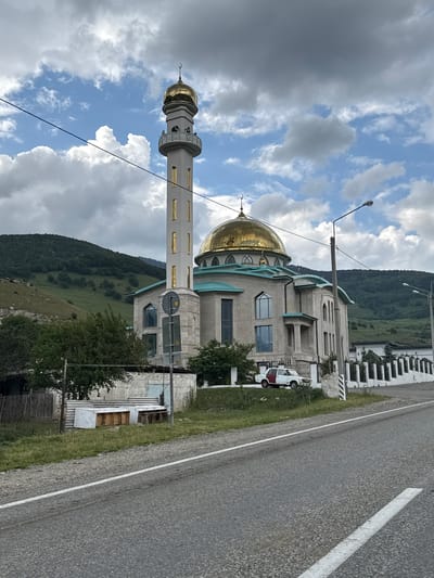 This media file captures a serene view of a mosque with a golden dome and a tall minaret. The mosque is a prominent structure, built with stone-like material and featuring elegant architectural details, including arched windows and decorative elements. Adjacent to the mosque, a white and red vehicle is parked, suggesting a peaceful, everyday scene. The setting is a mountainous region, with lush green hills and forested slopes forming a dramatic backdrop under a partly cloudy sky. The road, marked with a white line, runs alongside the mosque, indicating its proximity to a thoroughfare. A street lamp stands tall near the mosque. The overall atmosphere appears calm and picturesque, with the natural landscape complementing the man-made structure. The time of day appears to be daytime, with diffused sunlight filtering through the clouds, creating a soft illumination. There is no discernible text in the image.