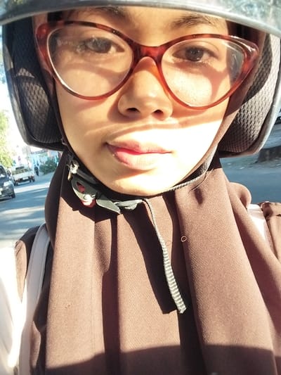 This is a portrait of a young person wearing a helmet and glasses, taken from a close-up perspective. The subject's face is partially obscured by the helmet and the large, red-rimmed glasses they are wearing. Their eyes are visible through the lenses, and their lips are slightly parted, colored with lipstick. The person is wearing a brown hooded garment. The photo appears to be taken outdoors on a sunny day, with strong shadows indicating it's likely daytime, possibly midday. The background shows a blurred street scene with vehicles, suggesting the person is in a public area, possibly in Macege, Indonesia, as indicated by the location context. The overall mood is casual and perhaps a bit informal, with the direct gaze through the glasses drawing attention to the subject's face. There is no visible text in the image.