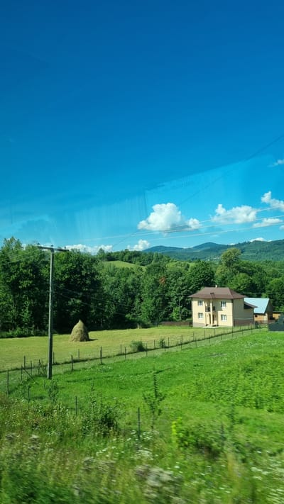 The image captures a rural landscape under a bright blue sky with scattered white clouds. In the foreground, a vibrant green meadow with wildflowers stretches across the frame. A wire fence runs horizontally through the middle ground, separating the meadow from a mowed field. To the left of this field, a tall, conical haystack stands prominently. A wooden utility pole with wires is also visible in this area.

Further into the background, a cluster of lush green trees forms a dense forest that extends to the foothills of rolling mountains. Nestled among the trees, on the right side of the image, are two houses. The larger house is light brown with a dark brown roof and multiple windows. Adjacent to it, a smaller building with a light blue roof is partially visible. The overall scene depicts a serene countryside setting. The location is identified as Krasnoilsk, Ukraine. There are no people or animals visible in the image, and no actions or interactions are depicted. The environment is natural and pastoral.