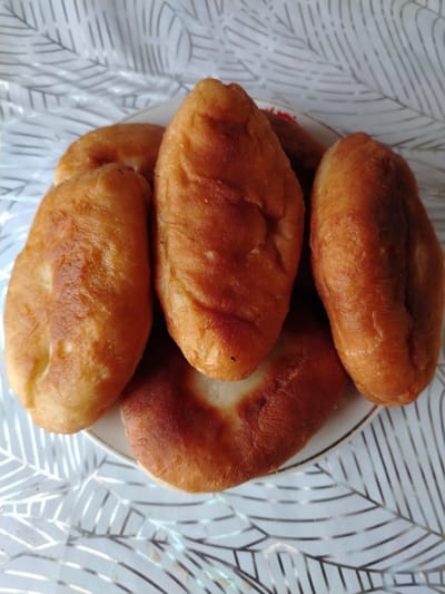 A close-up overhead shot shows a plate of golden-brown fried pastries resting on a white and silver patterned tablecloth. The pastries are oval-shaped with a slightly uneven surface texture, indicating they have been fried. They are arranged in a cluster on a white plate, with some overlapping. The tablecloth has a repeating pattern of stylized leaves or branches in silver on a white background. There are no people visible in the image, and no specific location in Жернов ауылдық округі, Kazakhstan, can be identified from the visual information provided. The scene appears to be a simple still life of food.