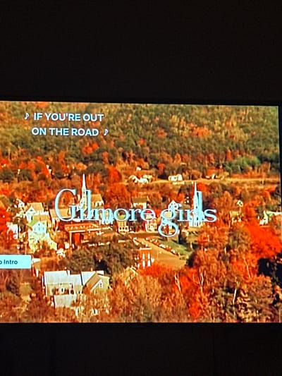 This image is a photograph of a display screen, likely a television, showing a still frame from a video. The video content depicts an aerial view of a small, rural town or village nestled within a dense forest, predominantly displaying autumn foliage with prominent red, orange, and brown hues interspersed with green. Numerous residential buildings and a distinct white church with a tall steeple are visible throughout the town. Winding roads are observable between the structures and trees.
Several text overlays appear on the screen:
- In the upper left corner, white text reads "♫ IF YOU'RE OUT ON THE ROAD ♫".
- Centered on the screen, large, stylized white text with a slight metallic sheen spells "Gilmore Girls".
- In the lower left corner, a light blue rectangular button with white text displays "Skip Intro".
The photograph itself was captured in the city of McAllen, United States of America. No individuals or specific actions are discernible within the displayed video content.