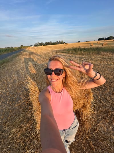 A blonde woman with sunglasses and a pink tank top is taking a selfie in a field of hay bales. She is smiling and holding up two fingers in a peace sign. The sun is setting, casting long shadows across the field. The location is specified as Trouville-Alliquerville, France, implying the image was taken in or around this area. The environment is rural, characterized by agricultural fields and a clear sky.