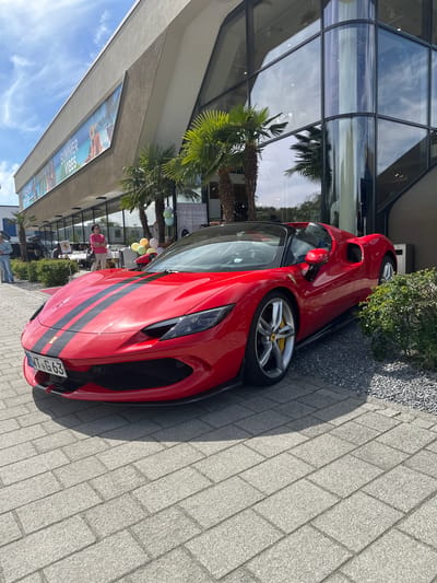A red Ferrari SF90 Stradale is parked on a gray brick sidewalk in Jestetten, Germany. The car has black racing stripes and a license plate that reads "WT-G 663". A modern building with a glass facade is behind the car, reflecting palm trees and the sky. There are people, including one seated woman in a pink shirt, near a tent or sheltered area. The sidewalk abuts a narrow strip of gravel and greenery. Part of a sign with the words "SUMMER VIBES" is visible on the building's awning.