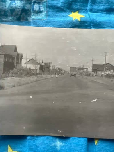 This is a black and white, vertically oriented photograph depicting a street scene in what appears to be a rural or suburban area, possibly from the mid-20th century. The foreground is dominated by a paved road and a line of buildings along the left side, suggesting a settled community. The buildings are of varying sizes and architectural styles, with some appearing to be houses and others possibly shops or businesses. Several utility poles with crossbars are visible along the street, indicating the presence of electricity or communication lines.
A few vehicles are present in the scene, including what looks like a truck driving down the road in the middle distance. The overall atmosphere is somewhat hazy, which could suggest either early morning, late afternoon light, or atmospheric conditions. There is no clear indication of specific weather, but the light suggests daytime. The surrounding area seems to have some vegetation, but it's not dense forest. The image is slightly faded and has some white specks, indicative of its age. There are no visible people clearly identifiable in the photograph. The background is a bright, overcast sky. The blue fabric with yellow stars on the left and right edges of the frame is not part of the original photograph but is part of the material the photograph is placed on, likely a tablecloth or blanket. There is no visible text within the photograph itself. The location context is provided as Дружба Снт, Russia.