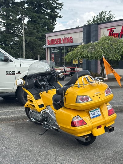 The image shows a yellow Honda motorcycle parked next to a white Ram 3500 truck. The motorcycle has an orange flag attached to it. In the background, there is a Burger King restaurant and a Tim Hortons. The scene appears to be in a commercial area. The weather is cloudy, and it is daytime. The license plate on the motorcycle reads "SAB 13". The Ram truck has "RAM 3500 HEAVY DUTY" written on the side. The Burger King sign is prominent, and some details about the address are visible: "19875-96 Ave. Bldg. A-Unit 1". The overall setting is an outdoor parking lot or street in front of businesses. There are no people visible in the image.