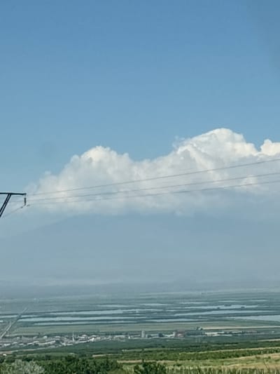 This media file shows a bright, clear blue sky with a large, fluffy white cloud dominating the left side of the frame. Several thin, dark power lines stretch horizontally across the image, intersecting with the cloud and extending towards the right. A single, dark power pole with insulators is visible at the bottom center of the frame, with supporting wires angled downwards.

The right side of the image depicts a landscape, appearing as a series of parallel horizontal lines that could represent agricultural fields or bodies of water, interspersed with green vegetation and some small structures that might be buildings. The overall scene suggests an outdoor setting, possibly rural, under a daytime sky. The lighting indicates it is daytime, with the sun likely high in the sky. There is no discernible text in the image. The image is oriented vertically, as if the camera was held upright.