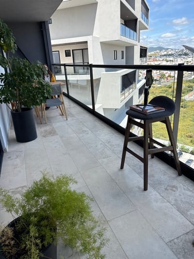 The image displays a modern residential balcony in Juriquilla, Mexico, offering an elevated view of the city, expansive undeveloped green terrain, and distant hills under a partly cloudy sky. The balcony floor consists of light-colored, rectangular tiles. A glass railing with dark metal top and bottom frames runs along the outer edge.
On the left, a tall potted plant in a dark gray cylindrical planter stands adjacent to a rectangular wooden table with two dark gray chairs. A person with dark hair, wearing a white top and carrying a yellow bag, is partially visible seated at one of the chairs. Closer to the foreground on the left, a second potted plant with light green, wispy foliage is situated in a dark container.
On the right side of the balcony, a dark gray upholstered stool with dark wooden legs is positioned near the railing. A closed book with a dark cover rests on the stool's seat. A black tripod stand with a vertically mounted smartphone is placed on top of the book, oriented to capture the exterior view beyond the railing.