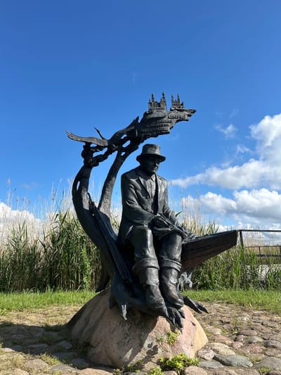 A bronze statue of a man sitting on what appears to be a wooden bench or plank is the central subject of the image. The man is wearing a hat, a suit jacket, and trousers tucked into boots. He is holding something in his hands, possibly a book or papers, and appears to be looking down at it. Behind him and to his left, a stylized bronze tree trunk curves upwards, supporting a depiction of a city skyline and what looks like a bird in flight. Text is inscribed on the city skyline portion, with the word "БЕЛАРУСЬ" clearly visible, indicating a connection to Belarus. The statue is positioned on a large rock, surrounded by cobblestones and grass. In the background, tall reeds or dry grasses grow, and beyond that, a wooden fence and a cloudy blue sky are visible. The scene is set outdoors, likely in a park or natural area, given the vegetation and open sky. The statue is located in the village of Azaretski, within the Azaretski Village Council, Belarus.