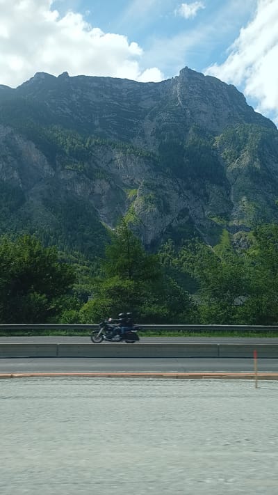 This image captures a mountainous landscape and a road in Golling an der Salzach, Austria. The upper two-thirds of the frame are dominated by a massive mountain range, characterized by steep, rugged grey rock faces interspersed with dense green coniferous forests. Lighter green vegetation patches are visible on some steeper mountain sections. Above the mountains, the sky is partly cloudy, with areas of clear blue visible between white cumulus clouds.
A multi-lane paved road runs horizontally across the mid-ground, separated by a grey metal guardrail and a concrete barrier. On the far side of this road, a dark-colored motorcycle is in motion, traveling from left to right. Two individuals, both wearing dark helmets, are riding the motorcycle. The immediate foreground consists of a light grey, textured surface, possibly gravel or an unfinished road. A slender wooden stake with a red tip is positioned vertically on the far right of this foreground. Motion blur is evident on the motorcycle and the immediate foreground, suggesting the photograph was taken from a moving vantage point.