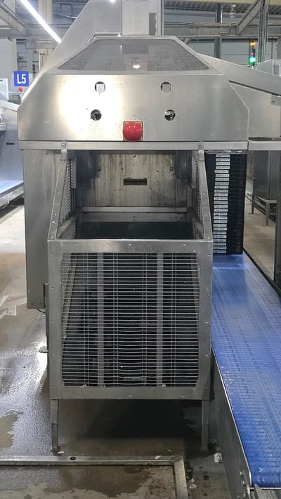 An industrial setting, likely a food processing facility in Crailsheim, Germany, is depicted. The foreground features a large, stainless-steel machine with a grated cage. Within the cage, a dark receptacle is visible. To the right, a blue conveyor belt runs parallel to the machine. In the background, a stack of black trays with labels is present. The machine has a prominent red emergency stop button and several circular openings on its surface. A blue sign with "L5" in white lettering is mounted on the left side of the machine. The floor appears wet and grimy, suggesting an operational environment. The lighting is artificial and bright, typical of an indoor industrial space. No individuals are present in the image.