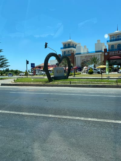 The image shows a traffic light positioned above a large, heart-shaped sculpture made of what appears to be tires, located in Alanya, Turkey. The sculpture is situated in a traffic island with greenery. Behind the sculpture, there are several multi-story buildings with modern architecture. One building has signage that reads "Aqua Fun City" and another has "GOD OF INK TATTOO & PIERCING". Several vehicles, including cars and a van, are visible on the road and in parking areas. The sky is clear and blue, indicating a sunny day. The overall scene depicts a street view in a commercial or entertainment area.