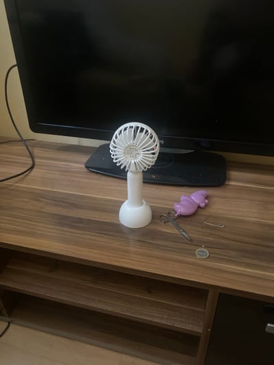 The image displays a wooden shelf unit with a dark television screen positioned above it. On the shelf, a white portable fan stands in the center. To the right of the fan, there are several small objects: a pair of scissors, a purple hippo-shaped toy, a safety pin, and a coin. The environment appears to be indoors, with a wall visible in the background. The overall scene suggests a personal space, possibly a living room or a desk area. There is no indication of a specific location within the city of Košice, Slovakia.