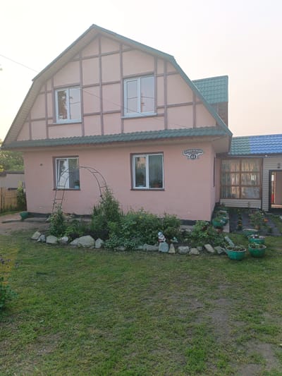 A pink, two-story house with a green, shingled roof stands in a yard. The house has white-framed windows and decorative half-timbering on the upper story. A smaller, attached sunroom with a blue-patterned roof extends from the side of the house. The sunroom has large windows with sheer curtains. The house is decorated with a sign that reads "Советская 61" (Sovetskaya 61). In front of the house, there is a garden bed with rocks, various plants, and small garden gnomes. Green planters filled with flowers are arranged in a line leading to the sunroom. A lawn in front of the garden is sparsely grassed in places, showing bare patches of dirt. An arched trellis stands in the yard near the house. The scene appears to be set in the late afternoon or early evening, with soft, diffused light suggesting overcast or hazy conditions. The setting is a residential yard, likely in a village or rural area, given the style of the house and landscaping. No people are visible in the image.