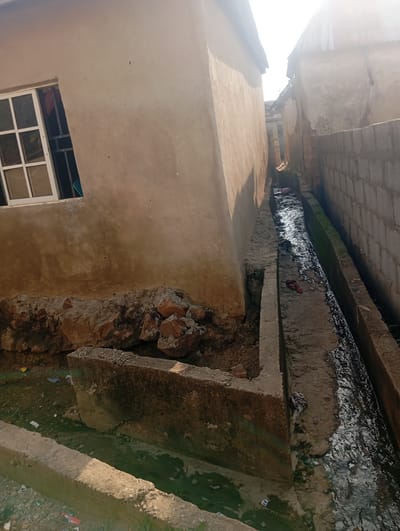 A high-angle, medium shot captures a narrow, unpaved alleyway in Gwagwa, Nigeria, flanked by two dilapidated buildings and a concrete drainage ditch running through the center. The building on the left features a pale, textured wall with a white-framed window and a metal security grate. On the right, a wall constructed of grey cinder blocks separates the alley from an adjacent property. The drainage ditch is filled with murky, dark water and debris, including what appears to be a piece of red fabric and a clump of grey fabric. Rocks and concrete rubble are piled near the base of the building on the left, adjacent to the drainage channel. The ground on either side of the ditch is littered with small pieces of trash, including wrappers and plastic. The sky is bright and hazy, suggesting either early morning or late afternoon. The overall impression is one of neglect and poor sanitation.