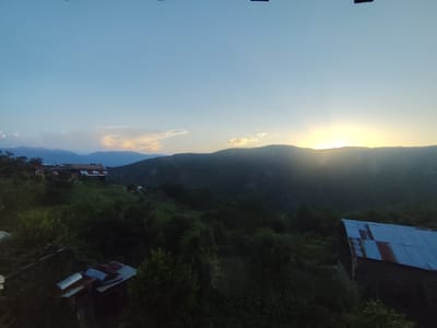 This media file depicts a rural landscape at dusk in Leshten, Bulgaria. The scene is dominated by rolling hills covered in lush green vegetation and trees. In the foreground, there are several rustic structures, including one with a corrugated metal roof showing signs of rust and age. A small, weathered building with a flat, metal-covered roof is also visible, along with some scattered debris.
In the mid-ground, a few houses are nestled on a hillside, their roofs also appearing to be made of traditional materials. The architecture suggests a traditional Bulgarian village setting. A road or path winds up the hill, with a couple of cars visible, hinting at the presence of inhabitants or visitors.
The background features a mountain range that fades into the distance, silhouetted against the sky. The sun is setting behind the mountains, casting a warm, golden glow and creating a dramatic and serene atmosphere. The sky transitions from a soft blue at the top to warmer hues of yellow and orange near the horizon, where a few wispy clouds are present.
The overall impression is one of tranquility and natural beauty. The time of day, indicated by the sunset, contributes to a peaceful and slightly melancholic mood. The location cues, such as the traditional architecture and mountainous terrain, strongly suggest a rural Bulgarian setting. There is no visible text in the image.