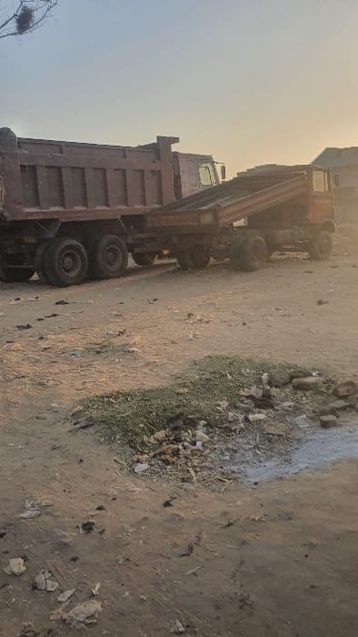A large, maroon dump truck is parked on a dirt and gravel surface. The truck's bed is raised. Debris and sparse vegetation are scattered on the ground near the truck. The sky, visible in the background, is hazy with sunlight. A small bird's nest is visible on a tree branch in the lower left corner.