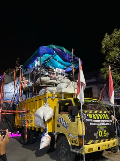 A modified yellow medium-duty truck is positioned on a street at night in Kedungbanteng, Indonesia. The truck's cab features a "CSS" logo on the door and "SPLaudio" and "NEXO" branding on the front bumper area. A prominent black and yellow banner, inscribed with "WARNING" and Indonesian text, including "05," is draped across the front. Two red and white Indonesian flags are mounted on poles at the front corners.

The truck's bed is heavily laden with sound system equipment, including large speakers and racks of electronics. A structure composed of metal scaffolding and wooden pallets supports a makeshift canopy made of blue and turquoise tarpaulins, covering some of the equipment. Numerous white sacks are stacked on the bed, with additional sacks hanging from the truck's side. Purple ambient lighting illuminates the base of the speaker setup on the left.

Four individuals are visible on or within the truck. One male stands on an elevated platform on the far left. Another male, wearing a striped shirt, is seated on a wooden platform on the truck bed. A third male stands near the flags on the right side of the truck bed, holding a bamboo pole. A fourth male occupies the driver's seat. In the lower-left foreground, a hand holds a smartphone, actively photographing the scene. The background reveals a dark night sky, a tree with green foliage on the right, and dimly lit buildings in the distance. The truck rests on an asphalt road.