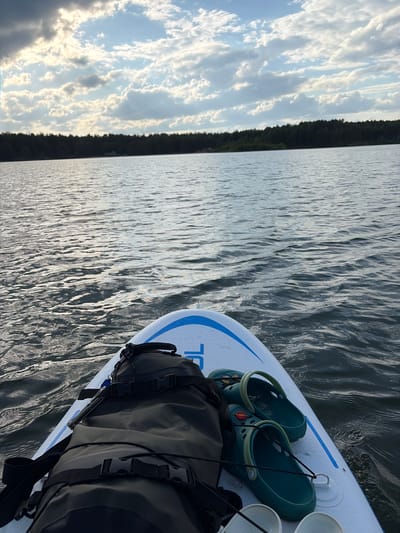 The image presents a view from a paddleboard on a body of water, likely a lake or river. The paddleboard is white with a blue stripe. On the board are a pair of green Crocs sandals and a dark-colored dry bag secured with black straps and buckles. The water exhibits gentle ripples and wavelets, reflecting the sunlight. In the background, a strip of land covered in dense trees is visible beneath a sky filled with scattered clouds. The overall setting suggests a calm, outdoor recreational activity. There is no information available in the image to suggest that this is located in Selychka, Russia.