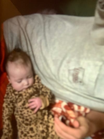 An indoor scene in Tulsa, United States of America, depicts an adult supporting an infant. The adult's upper torso and right arm are visible, clad in a light-colored t-shirt with a dark emblem on the left chest pocket area. The adult's right hand is partially visible, positioned near a patterned blanket. An infant, wearing a full-body leopard-print garment, lies horizontally across the adult's midsection with eyes closed, indicating sleep or rest. The infant's left hand rests on its chest, and dark hair is visible on its head. A red and white patterned blanket or cloth is draped over the adult's lower torso. A dark reddish-brown soft surface is visible adjacent to the infant's left side. The background is indistinct due to blur, suggesting a domestic interior.