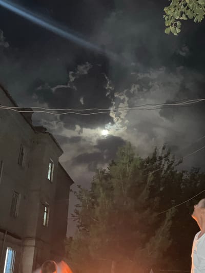 This is a low-angle, night-time photograph of a cloudy sky with the moon partially visible through the clouds. In the foreground, the top of a building with several windows is visible. Trees are silhouetted against the sky on the right side of the image. A bright, thin beam of light, possibly from a searchlight or spotlight, cuts diagonally across the sky on the left. Several electrical wires are also visible, running vertically and diagonally. A small cluster of green leaves from a tree is in the upper left corner. The image captures a somewhat dramatic and moody scene, with the bright moonlight and beam of light contrasting with the dark, cloudy sky and the shadowed building. There is no visible text in the image. The location context is Balkh, Tajikistan.