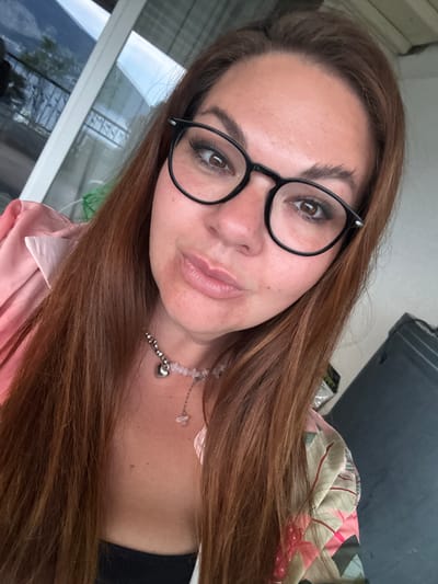 A selfie of a woman with brown hair, wearing black framed glasses and a necklace with a heart pendant. She is wearing a pink shirt. The background shows a window with a reflection of greenery and sky. A piece of fabric is draped in the background. The location cannot be verified based on the image.