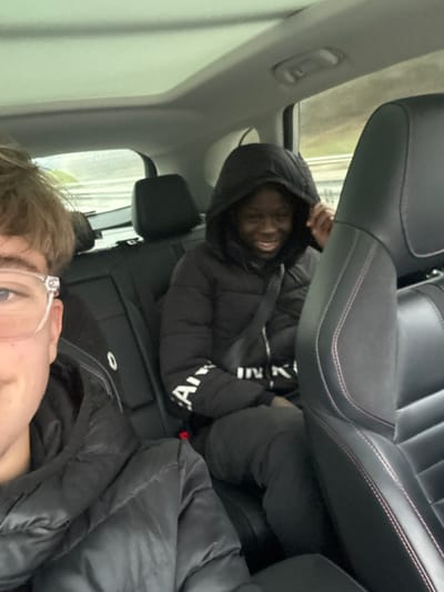 The image displays an interior view of a moving vehicle, reportedly in Eckartswiller, France. A person is partially visible on the left in the foreground, wearing a black puffer jacket and clear-framed glasses, with light brown hair, suggesting they are capturing the photograph. In the midground, seated in the rear right passenger seat, is an individual with dark skin, wearing a black hooded puffer jacket with the hood up. This person is smiling broadly towards the camera, showing teeth, and has their left hand raised near their head. A seatbelt is secured across their chest. The text "ARMANI" and "IN K" is partially legible on the right sleeve of their jacket. The car's interior features dark-colored seating, with visible red stitching on the front right seat. A sunroof is present in the ceiling. The environment outside the vehicle, seen through the windows, is blurred with tones of green and grey, indicating motion on a road or highway, with a guardrail faintly discernible.