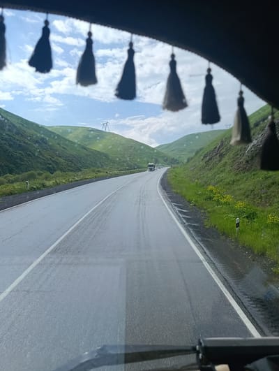 The image shows a view from inside a vehicle, looking out at a winding road through a lush green valley. The road is wet, suggesting recent rain, and reflects the overcast sky. Several dark, tassel-like ornaments hang from the top of the frame, partially obscuring the view of the sky and the upper parts of the rolling green hills.

The hills are covered in vibrant green grass and some sparse vegetation. Yellow wildflowers dot the grassy slopes along the side of the road. In the distance, a large truck and a few other vehicles are visible on the road. A prominent electrical pylon stands atop one of the distant hills, indicating the presence of power infrastructure in the area. The sky is cloudy with patches of blue peeking through. The overall impression is one of travel through a scenic, somewhat damp, natural landscape. There is no visible text in the image. The time of day appears to be daytime, likely late morning or afternoon given the lighting. The presence of the road and vehicles suggests a journey in progress.