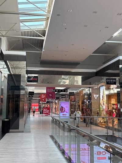 The image shows the interior of a shopping mall. A man walks down a brightly lit corridor lined with shops. The floor is made of pale wood-effect tiles. A glass balustrade runs along the right side of the corridor, overlooking a lower level of the mall. Above, a large skylight allows natural light to enter. Signs for various stores, including "Guess Kids", "Factory", "Salewa Kulig", and "Empik", are visible. A large red sign displaying "WY PRZE DAŻ%" and "-80%" suggests a sale. In the background, several people are browsing in shops. The scene depicts a typical commercial environment in a Polish shopping center.