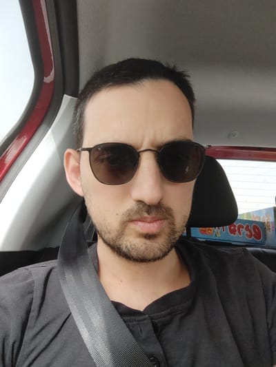 A man wearing sunglasses and a seatbelt is seated in a car. He has dark hair and a beard. The interior of the car is visible, including the ceiling and door frame. A package or box with colorful writing is partially visible near his seat. The exact location in Lod, Israel cannot be determined from this image alone.
