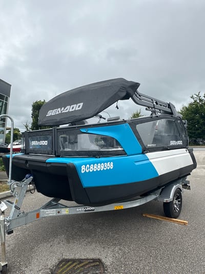 A Sea-Doo Switch pontoon boat, on a trailer, is parked on an asphalt surface. The boat is predominantly blue and white, with a black cover draped over part of it. The word "SEA-DOO" is visible in white text on the black cover. The registration number "BC8889358" is printed in white on the blue side of the boat. The trailer is silver and has a winch on the front. A wooden plank rests against the trailer wheel. The sky is overcast and appears to be daytime. The background includes some trees and hints of buildings. The scene suggests the boat is either being stored or is for sale. The location is the Township of Langley, Canada.