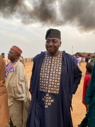 This media file captures an outdoor gathering in Agunu, Nigeria, likely during the daytime. The image is oriented vertically (rotated 90 degrees counter-clockwise), but if viewed upright, shows several individuals under a cloudy sky.
The main subject is a man standing prominently in the mid-right foreground. He is of African descent, smiling faintly and looking towards the camera. He wears a dark blue, traditional loose-fitting garment, possibly an agbada or caftan, featuring elaborate gold and silver square-patterned embroidery across the chest and down the front. He also wears a matching dark cap.
To his left and slightly behind him is another man, appearing older, also of African descent. He is dressed in a light beige or off-white traditional garment, adorned with a bold blue, red, and white geometric design on the chest. He wears a distinctive cap with a red base and a patterned brown or tan top. This man is looking downwards or to his left.
The background shows more people, some partially obscured, suggesting a crowd. Tents are visible in the distance, one appearing to have a flag. The ground is dry and light-colored, indicative of an arid or dusty environment. The sky above is largely overcast, with dense, dark clouds dominating the left side of the frame, possibly suggesting smoke, a dust haze, or an impending storm. No discernible text is visible on clothing or in the surroundings.