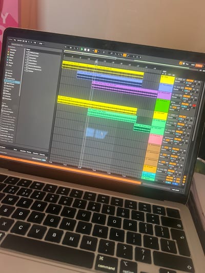 An open silver-colored laptop is depicted, its screen displaying the Ableton Live digital audio workstation interface. The software shows an arrangement view with multiple colored clips across various tracks, indicating an in-progress music production project. Track names such as "KICK," "FULL DRUM LOOP," "4 Serum 2," and "HAT LOOP" are visible on the right panel, along with mixer controls showing volume levels. The top bar of the software displays a tempo of 134.00 BPM. The laptop's black QWERTY keyboard with white lettering occupies the lower foreground. Reflections of an external light source are visible on the screen. The background is blurred, showing a light-colored surface. No individuals are present in the image. This scene is located in Winslow, United Kingdom.