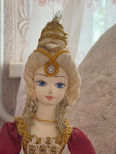 A close-up studio portrait shows a vintage doll with blonde hair styled in braids and an elaborate gold and jewel-encrusted headdress and costume. The doll has painted blue eyes, arched eyebrows, and red lips. The background consists of soft, blurred curtains and a textured fabric, suggesting an indoor setting. The overall scene is static and decorative, with no discernible action or interaction. The location is not specified in the image, but the user states it is in Lyambir', Russia.