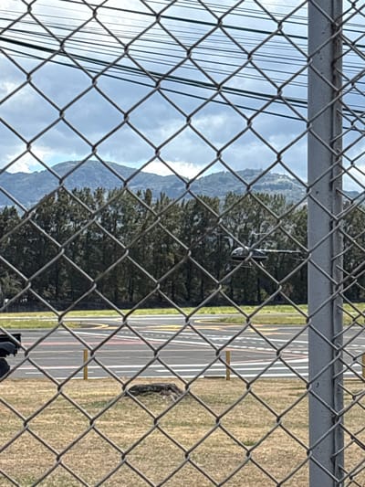 The image presents an outdoor scene viewed through a galvanized chain-link fence with a visible metal support post on the right. In the immediate foreground, a patch of dry, yellowish-brown grass covers the ground, featuring a dark, irregularly shaped object, possibly a tree stump or rock, in the lower-mid frame.

Beyond the fence and grass, an asphalt paved surface is visible, marked with distinct aviation symbols including yellow, red, and white lines, dashed markings, and circular patterns, consistent with a helipad or small airstrip. Several yellow bollards or ground markers are also present on the paved area. A dark-colored helicopter, appearing black or very dark blue, is partially visible above this paved surface in the midground, with its cockpit, fuselage, and tail boom discernible. Its main rotor blades are slightly blurred, suggesting either motion or a shallow depth of field. A dense line of tall, dark green coniferous trees forms a natural barrier behind the airfield.

In the background, a range of mountains stretches across the horizon, some densely forested, others appearing more rugged. One prominent mountain peak, visible towards the upper-right, is topped with several distinct radio or communication towers. The sky above is overcast, filled with various shades of grey and white clouds. Multiple dark power lines or utility cables run horizontally across the upper portion of the frame, behind the fence.

This scene is located within the San José metropolitan area of Costa Rica, specifically at or adjacent to the Tobías Bolaños International Airport (SYQ) in Pavas, San José, consistent with the aviation infrastructure and the distinctive mountainous backdrop, including peaks with communication towers common to the Central Valley.