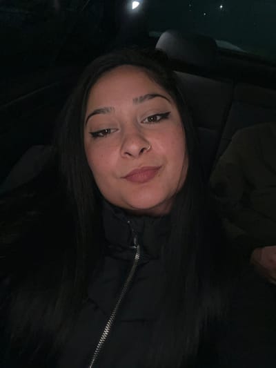 The image portrays a female individual seated within a vehicle during nighttime in the Gerakas Municipal Unit, Greece. The subject, positioned centrally, has long dark hair, dark eyes accented with winged eyeliner, visible freckles across her face, and a silver nose ring on her left nostril. She wears a black puffer jacket with a silver zipper extending down the front. Her gaze is directed towards the camera, and her lips display a slight upward curve. The background is dark and out of focus, suggesting low light conditions within the vehicle's interior. A faint circular light source is visible in the upper background. To the subject's left (viewer's right), the upper arm and shoulder of another individual wearing dark attire are partially visible, suggesting a second occupant in the vehicle. No direct interaction between the visible individuals is depicted.