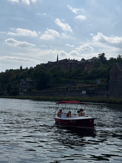 In Dinant, Belgium, a small red boat with a canopy is navigating the Sambre River. Four individuals are visible within the boat. The boat has the number "3" displayed prominently on its side, along with "INC8414305". In the background, a large, multi-story stone building with a spire dominates the hillside, likely a historical structure or institution. The river is calm with visible ripples, and lush green trees cover the slopes leading up to the buildings. The sky above is partly cloudy with bright white clouds against a blue backdrop.