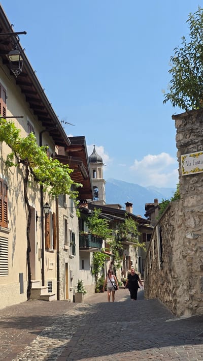 In Limone sul Garda, Italy, a narrow, cobbled street slopes upwards between tightly packed buildings. The facades of the buildings are a warm, pale yellow, with traditional wooden shutters and tiled roofs. Lush green vines are trained along some of the walls, adding a touch of vibrant nature to the scene. In the distance, a bell tower with a dark, domed roof rises above the rooftops, and beyond that, hazy blue mountains form the backdrop under a clear sky with scattered white clouds.

Two individuals are ascending the street. On the left, a woman with dark, shoulder-length hair wears a white tank top and light-colored shorts. She carries a dark crossbody bag. She is looking towards the camera with a slight smile. To her right and slightly behind her, a person with dark hair wears a black t-shirt and dark pants. They are holding a white paper in their right hand and appear to be looking downwards. Both are walking uphill on the cobblestone path.

A stone wall with a sign that reads "Via Fontanella" decorated with yellow lemons is visible on the right side of the street, adding to the distinctive charm of the Italian locale. Outdoor lighting fixtures are mounted on the buildings, hinting at the street's appearance after dark. The overall impression is one of a picturesque, historic Italian village on a sunny day.