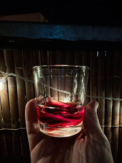 A hand holds a clear glass containing a red liquid, possibly wine or a cocktail. The glass is illuminated by string lights wrapped around it and behind it, creating a warm glow. The background consists of a woven bamboo fence, typical of outdoor patios or garden decorations. Above the fence, a dark blue illuminated surface is visible. The scene appears to be set outdoors at night. The specific location in Sinzheim, Germany, is not discernable from the visual information provided.